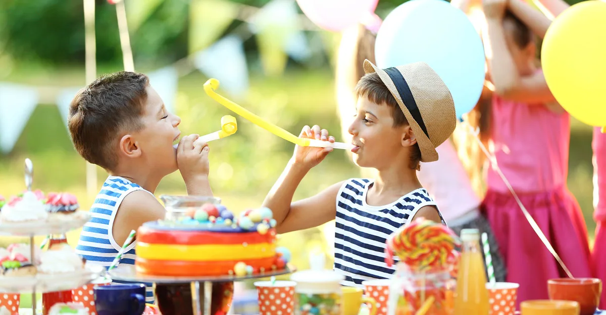 Two boys in striped shirts blowing party horns at an outdoor birthday party with colorful balloons and rainbow cake.