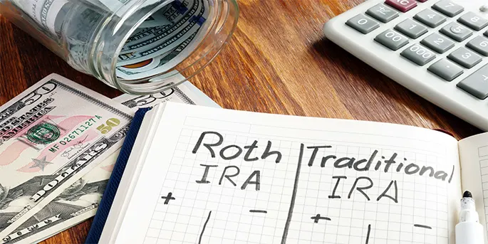 Notebook comparing Roth IRA and Traditional IRA with cash in jar and calculator on wooden desk