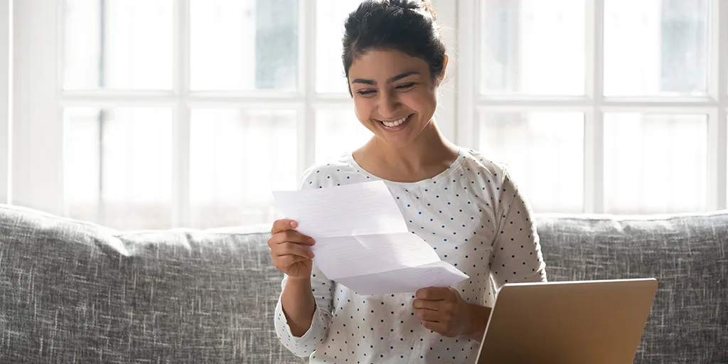 Smiling woman in polka dot shirt reading a document while sitting on a gray couch near bright windows.