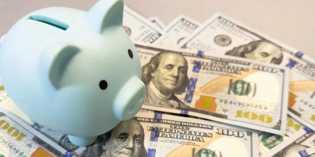 Light blue piggy bank sitting on scattered one hundred dollar bills on a beige surface.