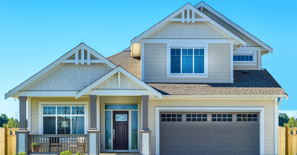 Two-story craftsman-style home with beige siding, white trim, peaked gables, front porch, and two-car garage against blue sky.