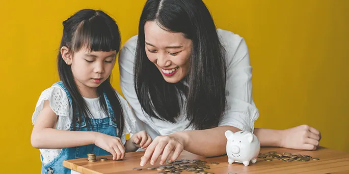 Mother and daughter counting coins at a wooden table with a white piggy bank against a yellow background.