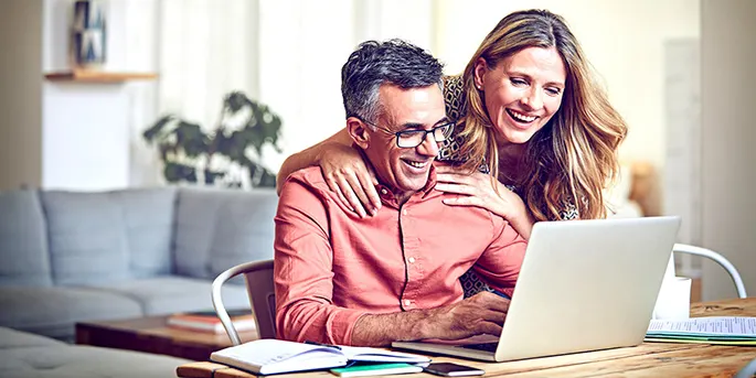 Smiling couple looking at laptop together at home, woman standing behind seated man in bright modern living room