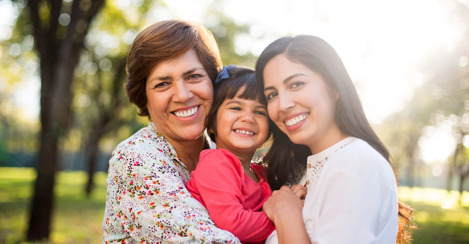 Three generations of smiling women embracing outdoors in a sunny park with trees in the background.