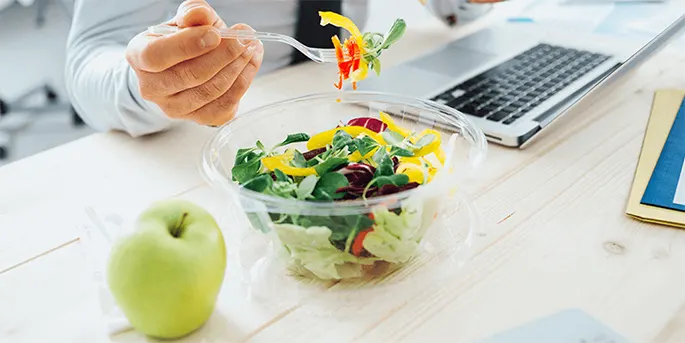 Person eating fresh colorful salad from clear bowl at desk with laptop, green apple, and notebook nearby.