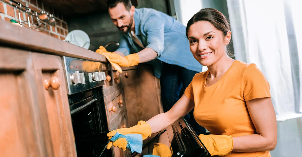 Woman in orange shirt and yellow gloves cleaning oven, smiling at camera, while man in blue shirt cleans in background.