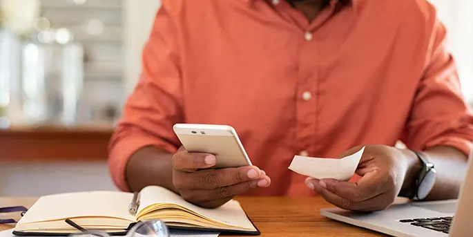 Person in coral shirt holding smartphone and receipt at desk with open book and laptop in bright office setting.