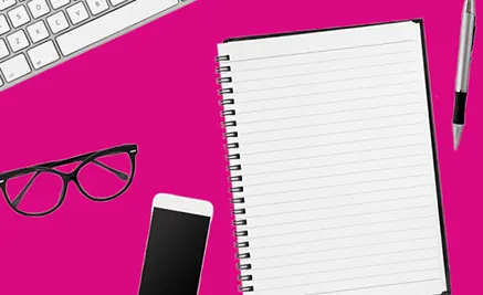 Flat lay of open spiral notebook, smartphone, black eyeglasses, keyboard, and pen on bright pink desk surface.