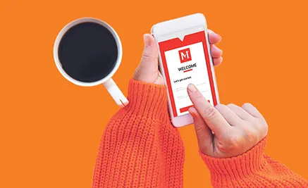 Hand in coral sweater holding white smartphone displaying app mockup, with black coffee cup on orange background