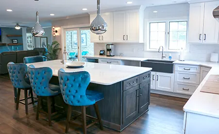 Modern kitchen with white countertops, dark island with blue tufted chairs, white cabinets, and stainless steel pendant lights.