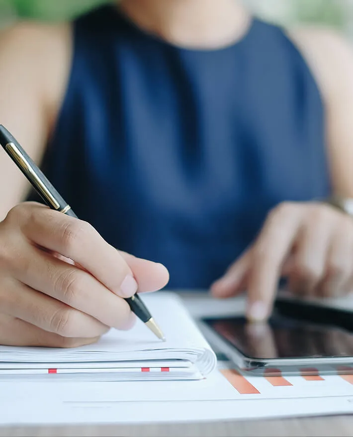 Person in navy blue sleeveless top writing in notebook with pen while using tablet device on desk