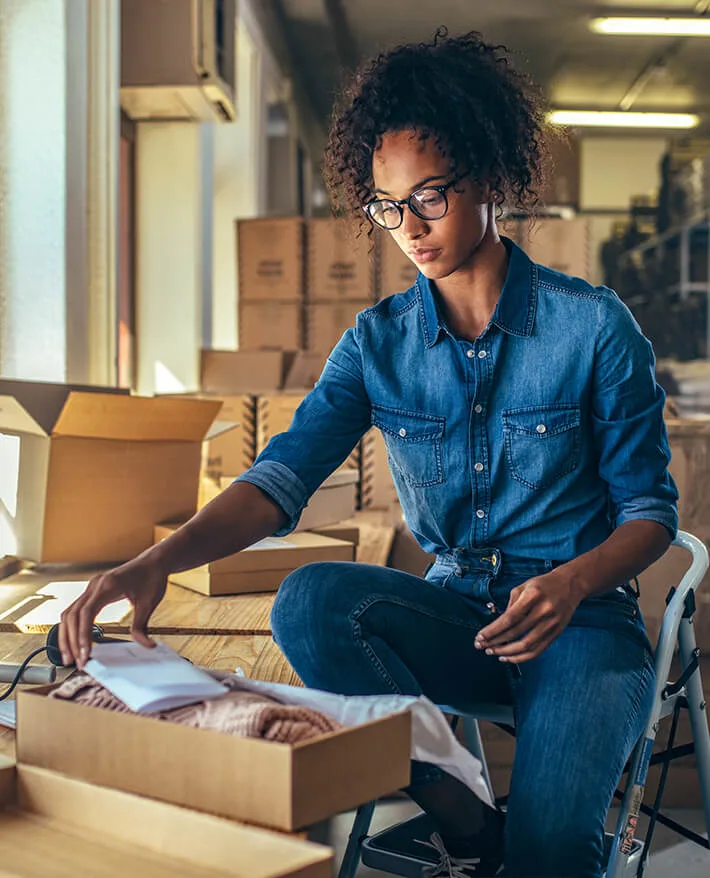 Woman in denim shirt and glasses packing cardboard boxes in a warehouse with stacked boxes in the background.