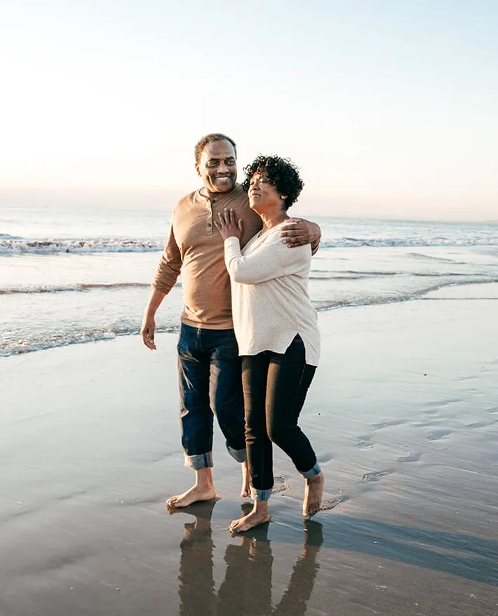 Happy couple walking barefoot on beach at sunset, embracing and smiling, with ocean waves in the background.