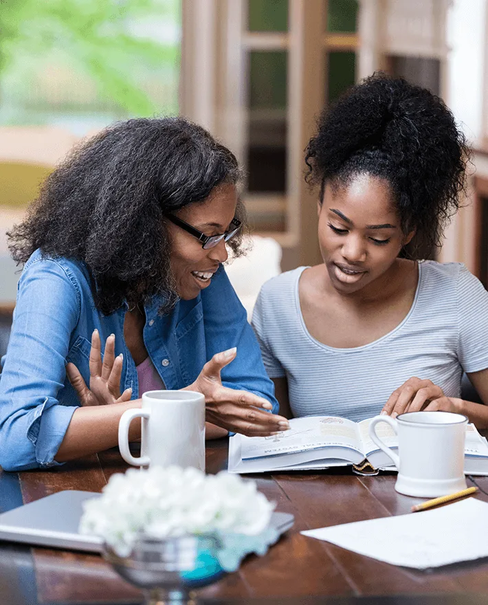 Two women studying together at a table with coffee mugs and an open book, engaged in conversation in a bright room.