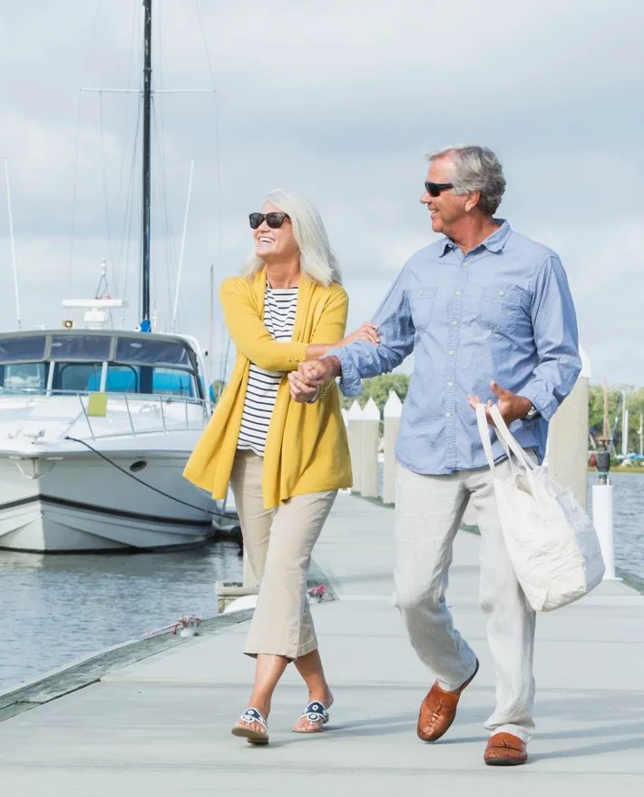 Senior couple walking hand in hand on marina dock with sailboats in background, wearing sunglasses and casual attire.