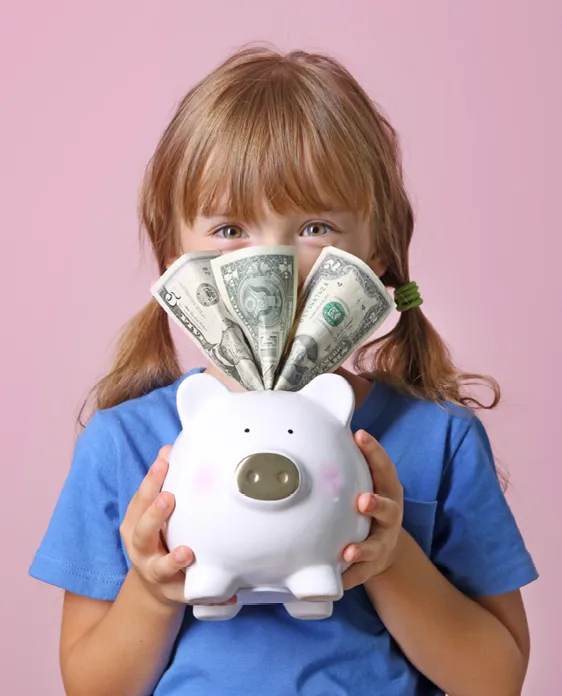Child in blue shirt holding white piggy bank with dollar bills fanned out on top against pink background