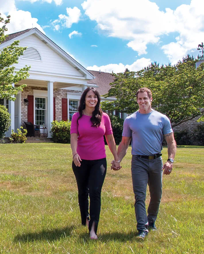 Happy couple holding hands walking on lawn in front of white brick house with red shutters under blue sky with clouds
