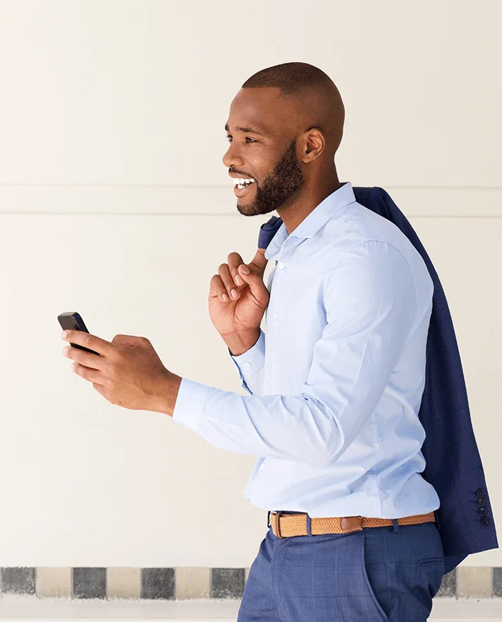 Smiling businessman in light blue shirt holding smartphone and navy blazer over shoulder against beige wall