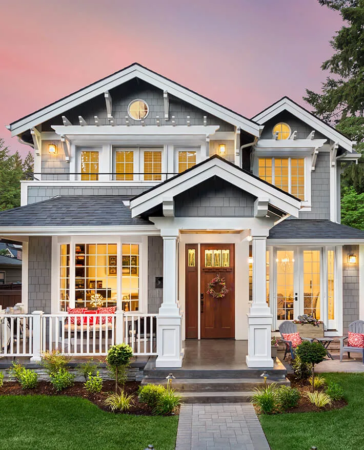 Gray craftsman-style house with white trim and glowing windows at dusk, featuring a covered front porch and pink sky.