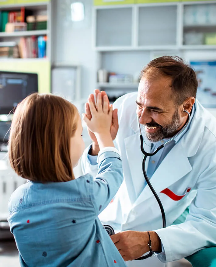 Smiling doctor in white coat giving high-five to young patient in medical office with shelves in background.