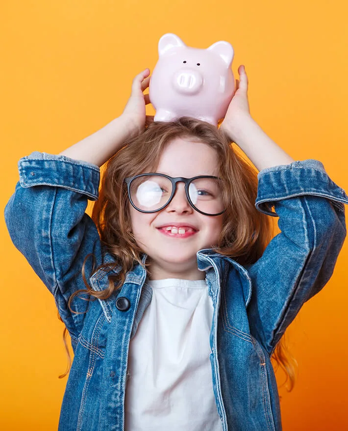 Smiling child wearing oversized glasses and denim jacket holds pink piggy bank above head against yellow background