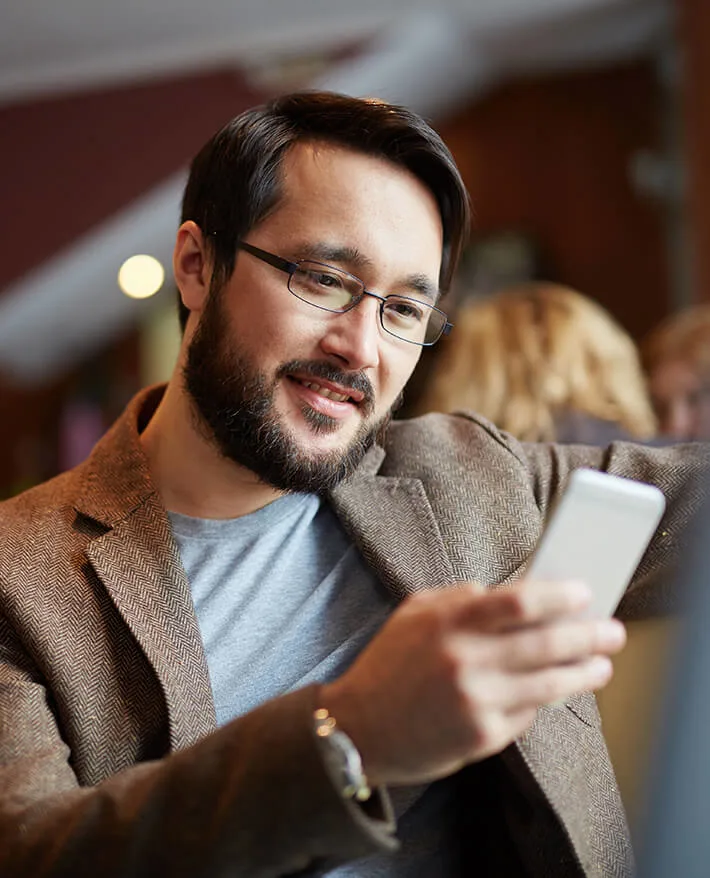 Smiling man with glasses and beard wearing brown blazer looking at smartphone in casual indoor setting