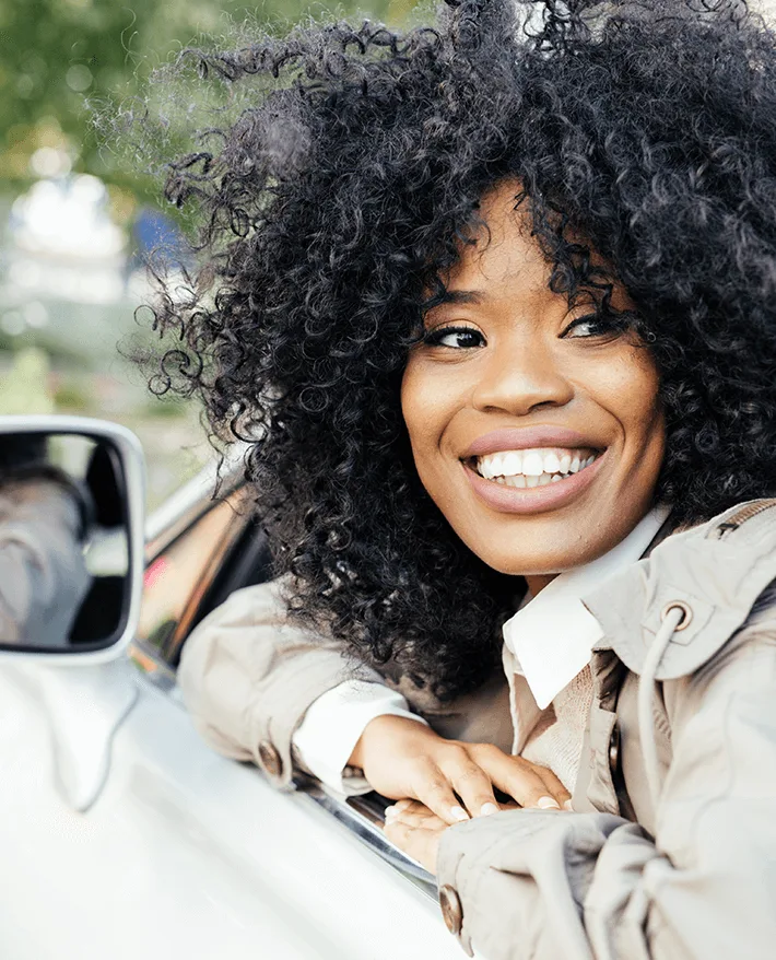 Smiling woman with curly black hair leaning out of white car window wearing beige jacket on sunny day with blurred background