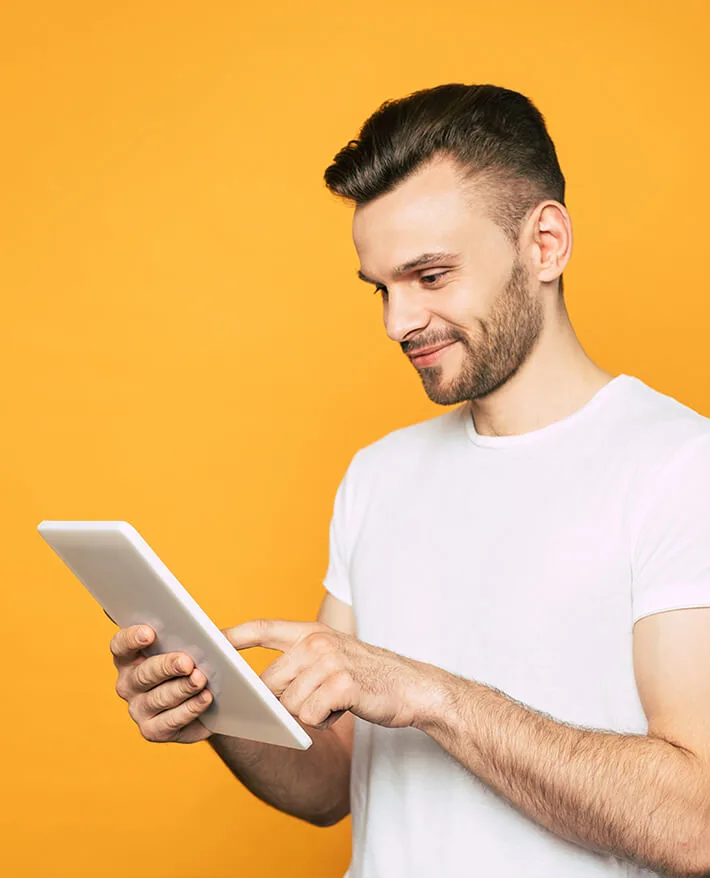 Young man in white t-shirt smiling while using a tablet against a bright orange background.