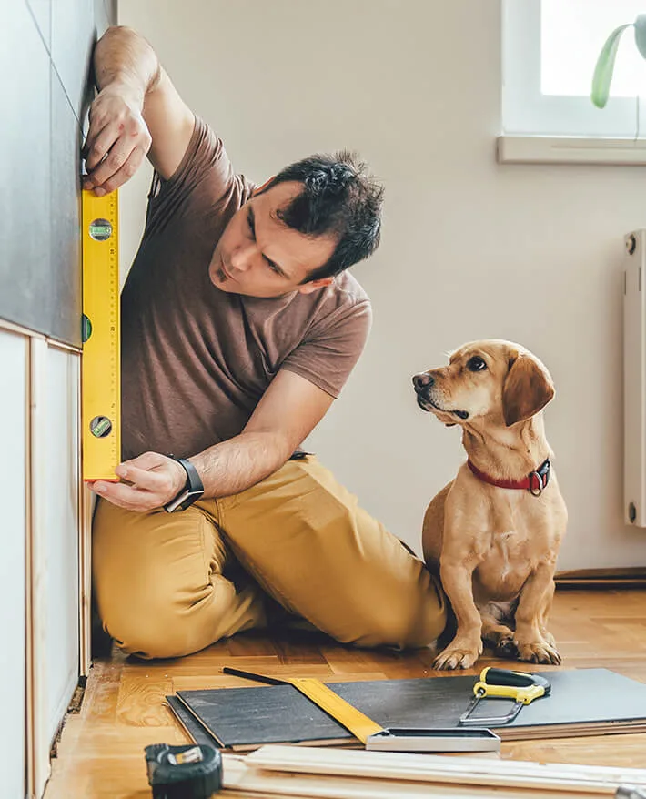 Man in brown shirt using yellow level on wall while tan dog with red collar watches during home renovation project.