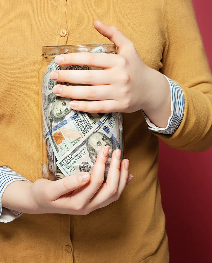 Person in yellow cardigan holding a glass jar filled with U.S. dollar bills against their chest.