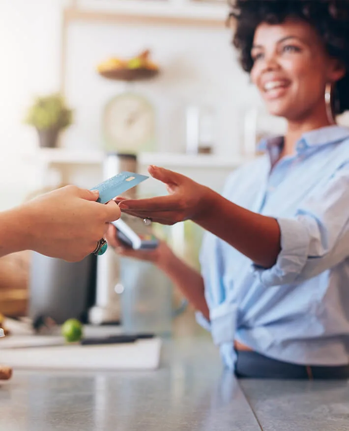 Smiling woman in blue shirt receiving a credit card from another person in a bright kitchen setting.