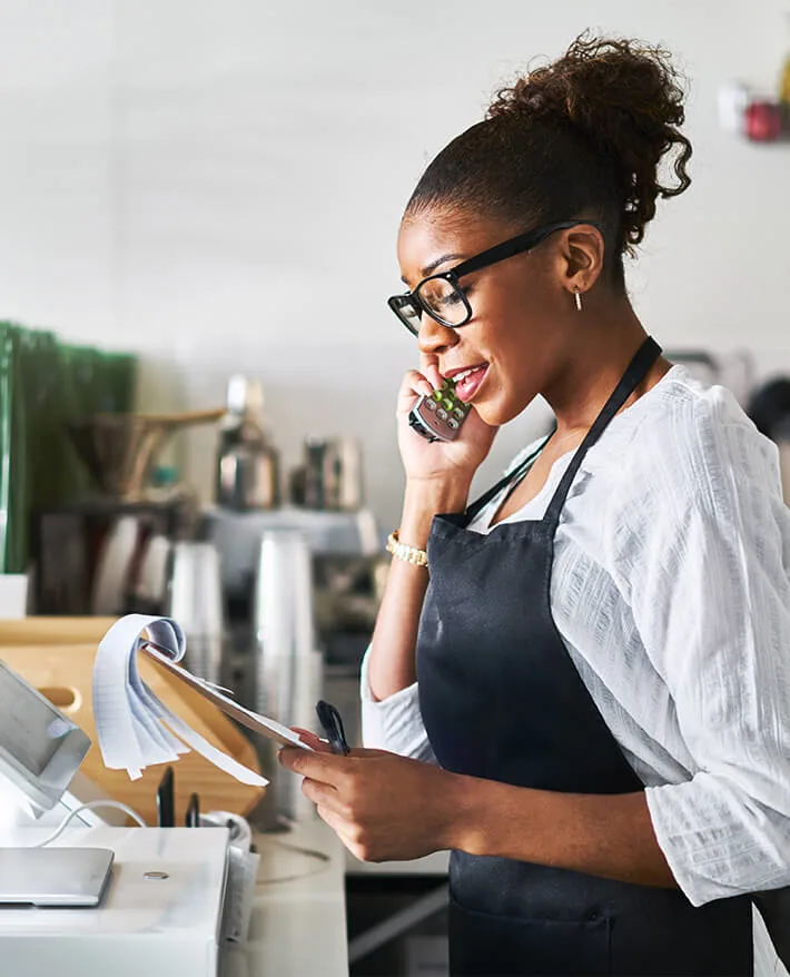 Small business owner in black apron and glasses talking on phone while reviewing receipts at cash register counter
