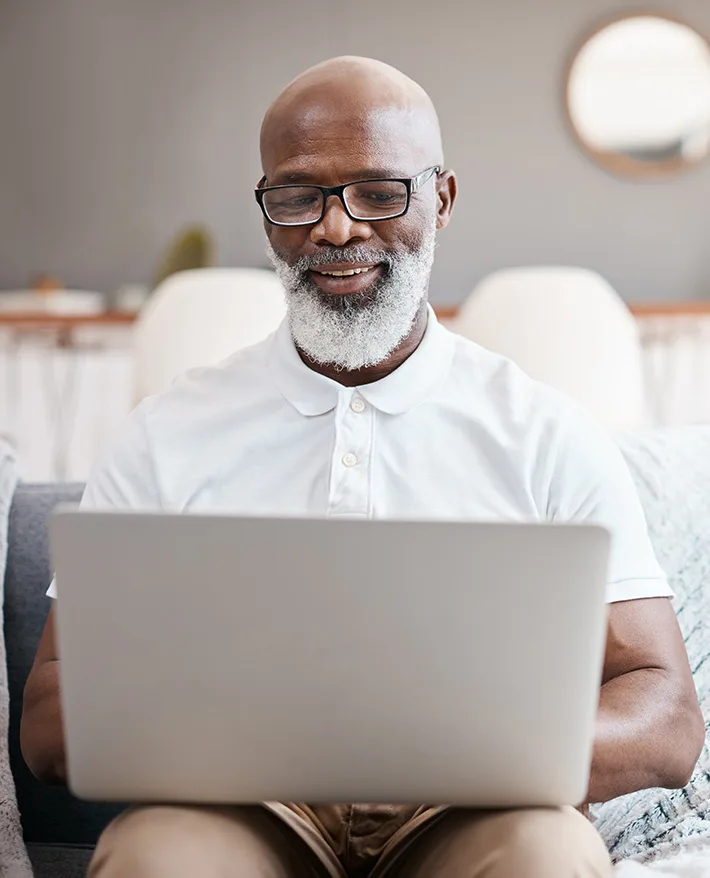 Smiling bald man with white beard and glasses wearing white polo shirt using laptop computer in modern home interior