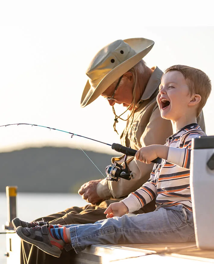 Young boy in striped shirt laughing while fishing with adult wearing sun hat on boat dock at sunset over water.