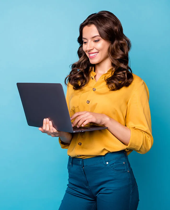 Smiling woman with wavy brown hair in yellow shirt holding and typing on laptop against blue background