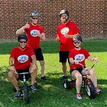 Four children in red shirts pose with tricycles on grass in front of a brick building