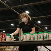 Volunteer in black shirt organizing food boxes on shelves in warehouse with industrial ceiling and lighting