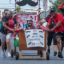 Group in red and black shirts marching in parade with mustache-themed banner and decorations