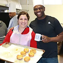 Woman in white apron with red collar holds tray of cupcakes next to smiling man with coffee mug in kitchen setting
