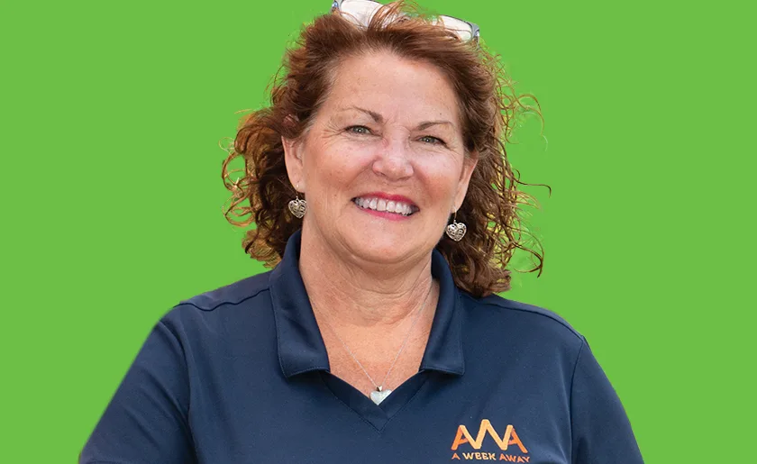 Woman with curly auburn hair smiling in navy blue polo shirt with orange logo against bright green background.
