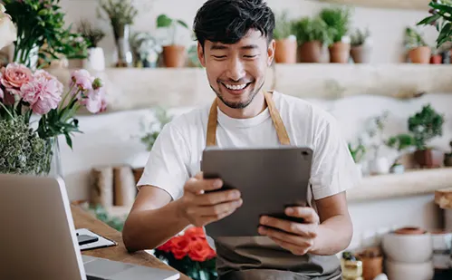 Smiling male florist in apron using tablet in flower shop with plants and laptop on counter