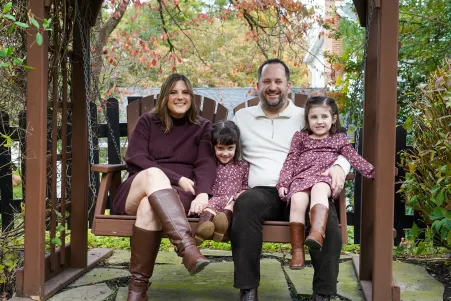 Family of four sitting on wooden swing in autumn garden, parents with two daughters in coordinating burgundy outfits.