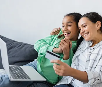 Two smiling women sitting together on a couch, looking at a laptop while one holds a credit card for online shopping.