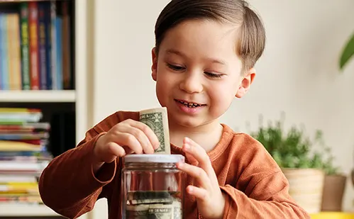 Young boy in orange shirt smiling while putting dollar bill into clear glass jar, with bookshelf in background