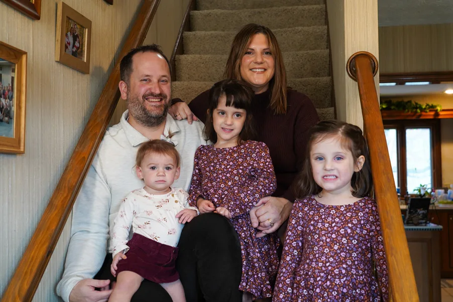 A smiling family of five sits together on carpeted stairs in their home, with parents and three young children posing for a photo.
