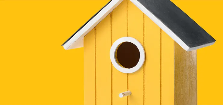 Bright yellow wooden birdhouse with white trim, circular entrance hole, and dark gray roof against yellow background.