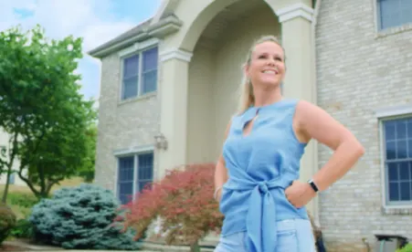 Woman in blue dress standing confidently in front of a two-story brick home with landscaped yard and trees.