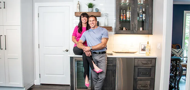 Smiling couple posing in modern kitchen with gray cabinets, white backsplash, and open shelving with liquor bottles.