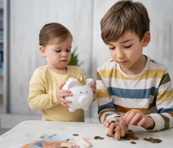 Two children counting coins at a table, one holding a white piggy bank while the other stacks coins.