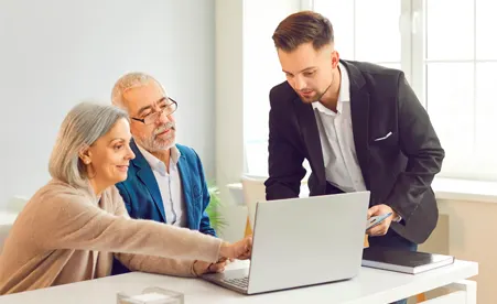 Young businessman in black suit presenting on laptop to older couple in bright modern office setting